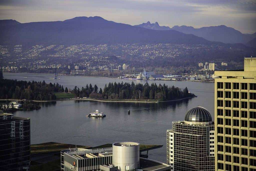 View of Vancouver with tall buildings in the foreground and a small forested island sitting in the harbor with mountains in the distance. The coastal scenery and winding waterfront roads reflect the landscapes travelers encounter on scenic road trips in British Columbia.