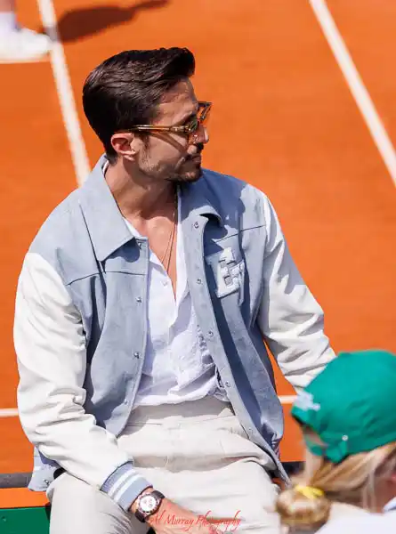 A stylish man wearing sunglasses and a light blue jacket sits courtside at the Monte Carlo Masters, with clay tennis courts and a uniformed event staff member in the background. Captures the atmosphere and seating proximity described in a Monte Carlo Masters seating guide.