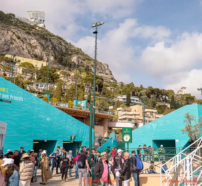 Crowds gather at the Monte Carlo Masters entrance, surrounded by turquoise walls, signage, and a scenic hillside dotted with buildings and greenery. A snapshot that helps illustrate accessibility and atmosphere in a Monte Carlo Masters travel guide for seniors.
