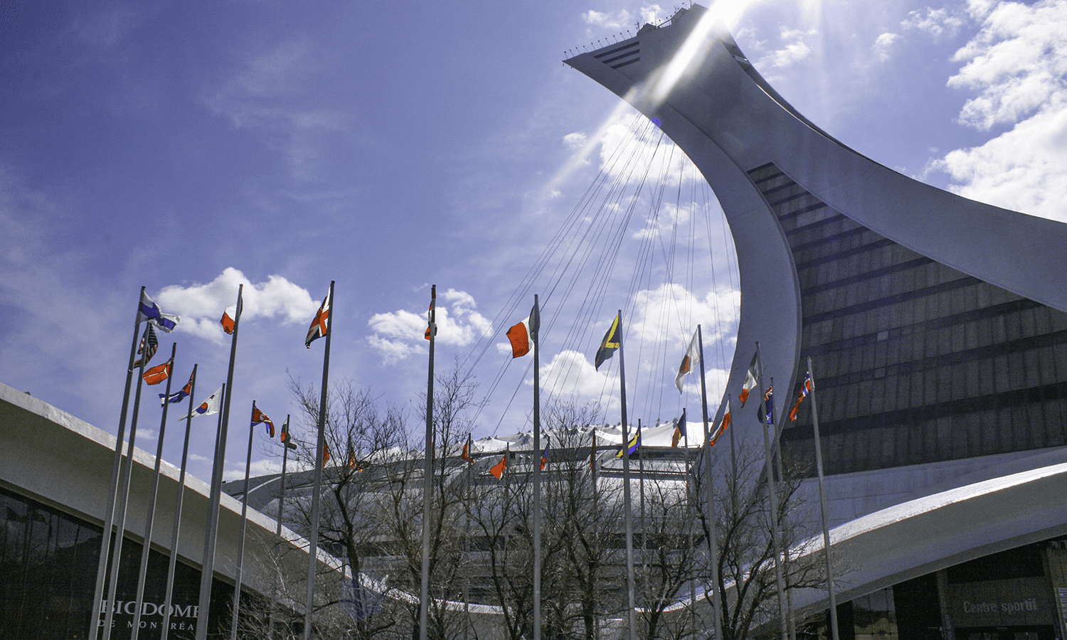 Modern view of Montreal&rsquo;s Olympic Stadium under a bright spring sky, with international flags fluttering in the foreground. A popular landmark often featured in guides on what to do in Montreal in spring.