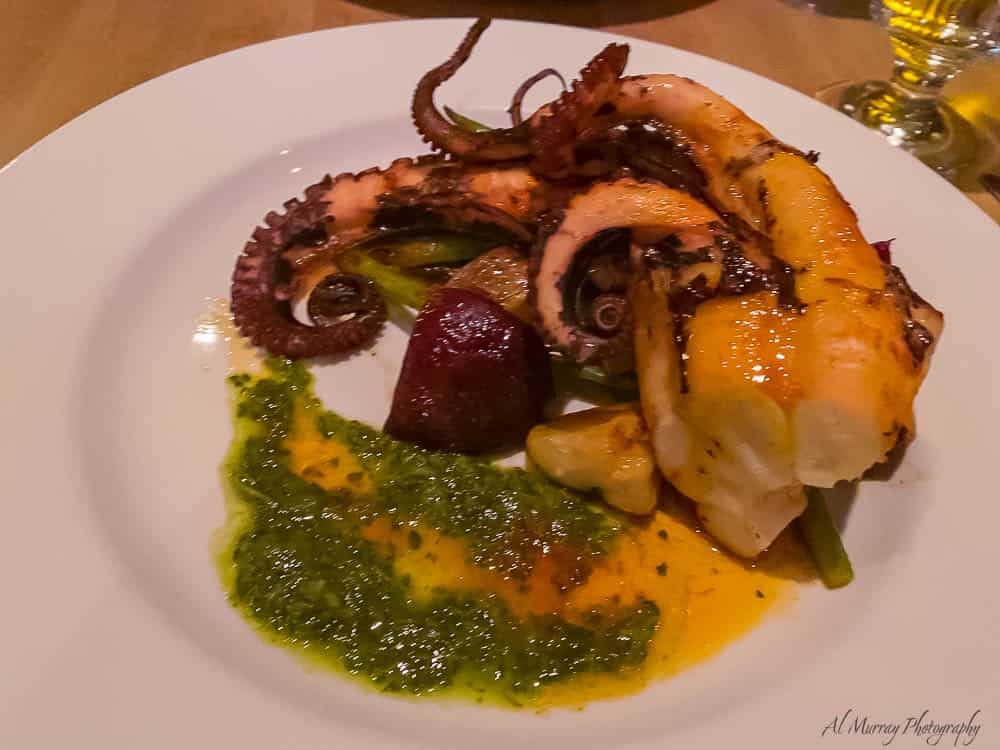Close-up of a gourmet seafood dish featuring grilled octopus, roasted vegetables, and a vibrant green herb sauce, served on a white plate at a Montreal restaurant. Sampling seasonal cuisine is one of the top things to do in Montreal in spring for food lovers.
