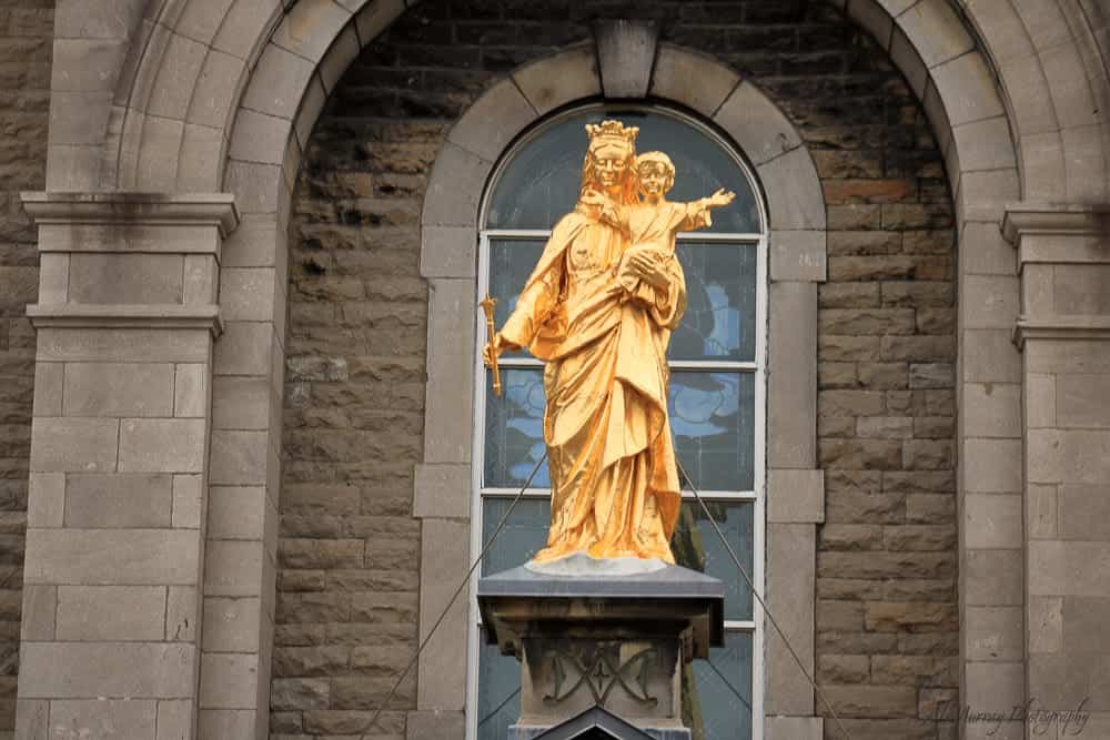  Golden statue of the Virgin Mary holding baby Jesus, mounted above the entrance of a historic stone church in Montreal. Visiting sacred landmarks like this is a peaceful and reflective option for what to do in Montreal in spring.
