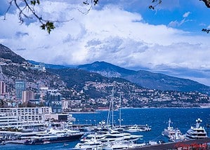 A panoramic view of Monte Carlo’s coastal cityscape backed by mountains under a cloudy sky, with the Mediterranean Sea in the foreground. The hilly terrain shown underscores the importance of senior mobility tips for visiting Monte Carlo safely and comfortably.