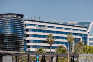 Exterior view of the sleek, modern Sheraton Hotel at Nice Airport, featuring curved glass architecture and surrounded by palm trees under a clear blue sky. A key location for travelers researching a Nice Airport Sheraton review, offering convenience and upscale amenities steps from the terminal.