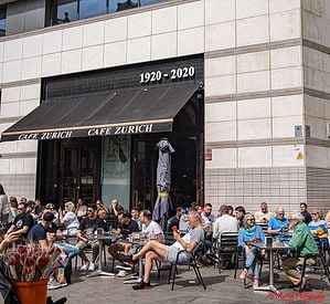 Seniors and other patrons relax at outdoor tables of Café Zurich in Barcelona, beneath a black awning marked “1920–2020.” This centrally located café offers a comfortable rest stop for seniors exploring the city during the Barcelona ATP Tennis tournament.