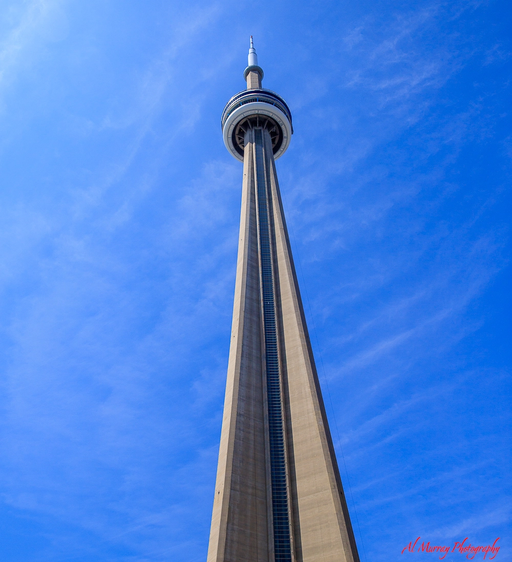 View looking up at the CN Tower in Toronto against a bright blue sky, highlighting the 360 Restaurant's circular observation deck near the top. The image conveys the dramatic height and setting for a unique dining experience at 360 Restaurant CN Tower Toronto.