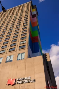 Upward view of the Hyatt Place Montreal building featuring a beige high-rise with a vertical rainbow mural on one side and the Banque Nationale sign at the base. The striking exterior sets the scene for a Hyatt Place Montreal review for senior travellers looking for centrally located accommodations.
