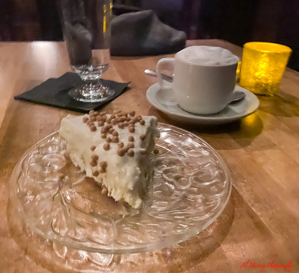 Slice of creamy cheesecake topped with small caramel bits served on a clear glass plate beside a cappuccino with foamed milk and a glowing candle on a wooden table. The cozy dessert and coffee pairing highlights a sweet finish featured on the restaurant L Orignal menu.