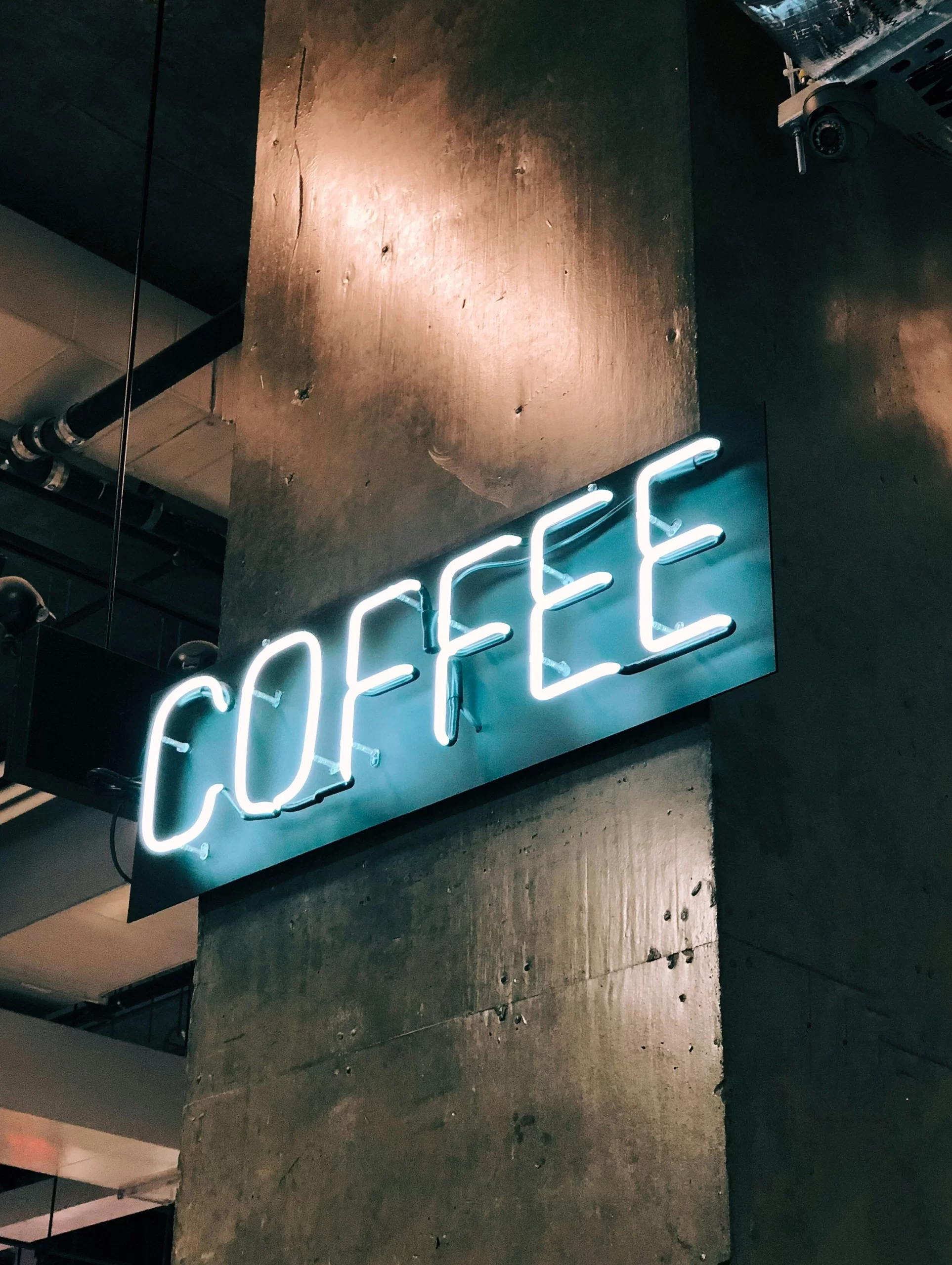Neon sign reading “COFFEE” in bright blue letters, mounted on a concrete pillar inside an industrial-style café with exposed pipes and ducts. The modern interior suggests a trendy spot popular among travellers looking for the best coffee shops in Montreal.