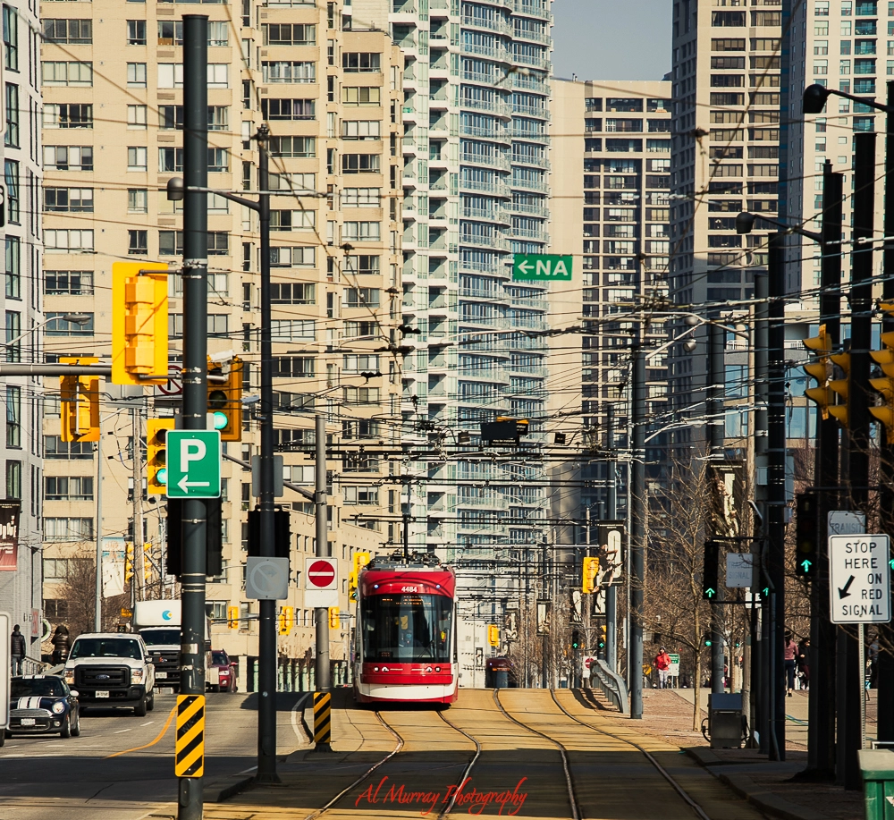 A red and white Toronto streetcar travels down a track-lined street surrounded by tall condo buildings and traffic lights, capturing an urban cityscape ideal for budget sightseeing. The dense overhead wires and visible street signs emphasize public transit access in downtown Toronto.