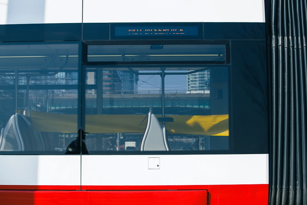 Close-up view of a modern Toronto streetcar window showing empty grey seats and a yellow barrier, with city buildings and an overpass reflected in the glass. Part of a Toronto streetcar sightseeing tour route, the streetcar’s LED display shows unreadable destination text