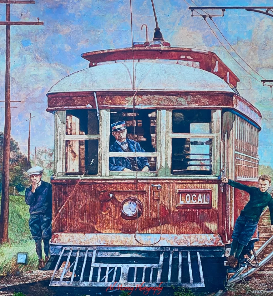 A vintage-style mural depicting a red and white streetcar labeled “LOCAL,” with a uniformed conductor at the window and two young boys—one leaning against the car and the other hanging onto the side. The artwork, painted with textured brushstrokes, is part of an Irish pub Etobicoke, celebrating historic street scenes and local heritage. Overhead wires and wooden utility poles add to the nostalgic setting.