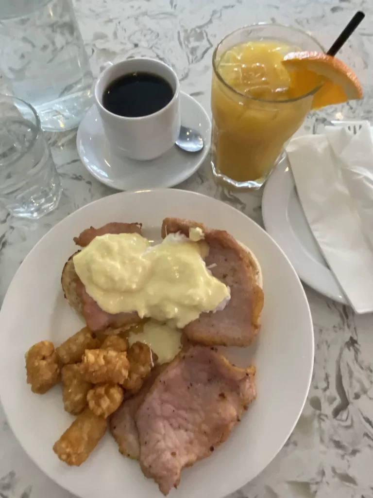 Breakfast plate at an Italian cafe in Toronto’s Little Italy featuring peameal bacon, crispy tater tots, and eggs topped with creamy hollandaise sauce. Served with a glass of orange juice garnished with an orange slice and a cup of black coffee. A hearty option at one of the best Italian cafes in Little Italy Toronto.