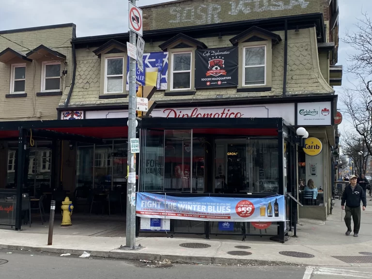 Exterior of Café Diplomatico in Toronto featuring a covered patio with promotional signage for a winter dining deal and a banner reading “Soccer Headquarters” above. A blue banner advertises “2 can dine for $59” alongside drink images, reinforcing its role as a local hangout. Ideal image for a “Cafe Diplomatico Toronto review.”
