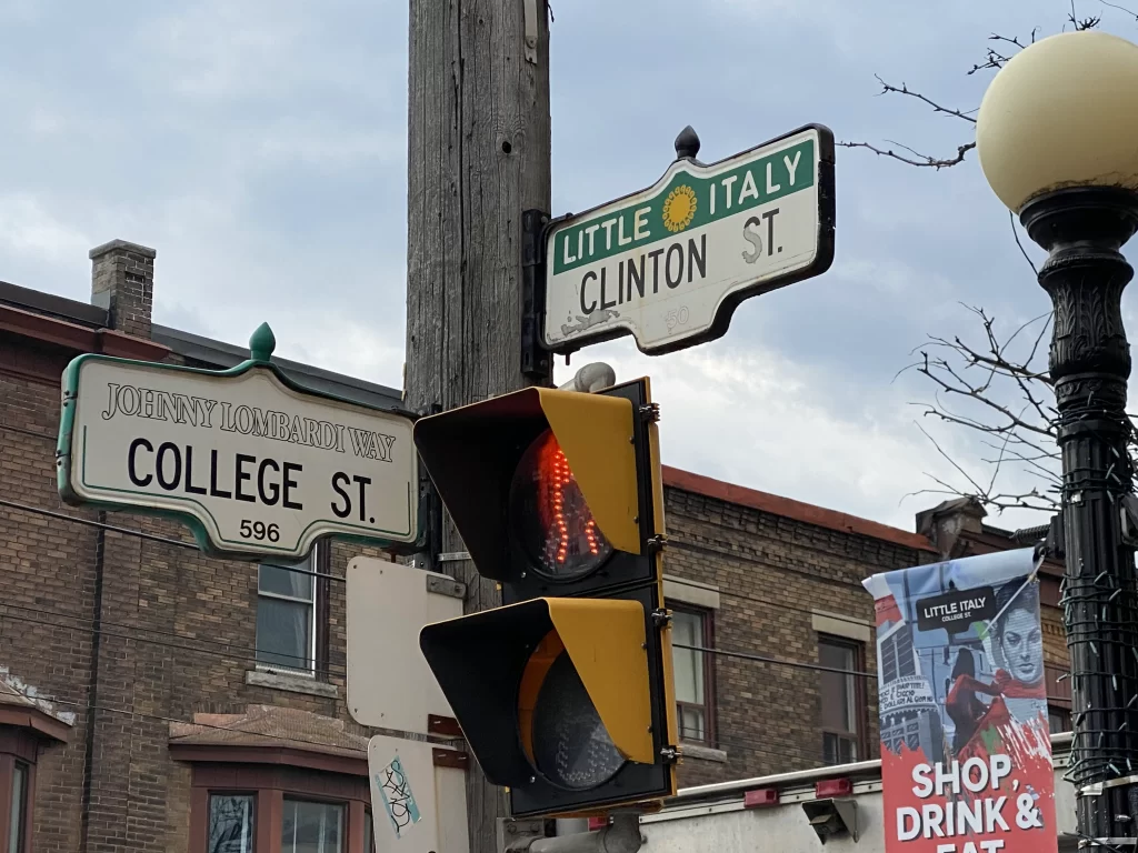 Street signs for College Street and Clinton Street in Toronto’s Little Italy, with College also labeled as Johnny Lombardi Way. A nearby banner promotes the area with bold red text reading “Shop, Drink & Eat” under the Little Italy College St branding. Sets the scene for exploring the best Italian cafes in Little Italy Toronto.