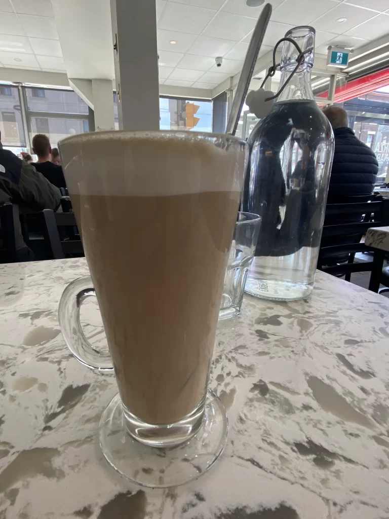 Close-up of a tall glass latte with a thick foam layer, served on a marble table alongside a glass water bottle in a bright, bustling cafe. Large windows in the background fill the space with natural light and overlook a street corner in Little Italy. A cozy moment at one of the best Italian cafes in Little Italy Toronto.