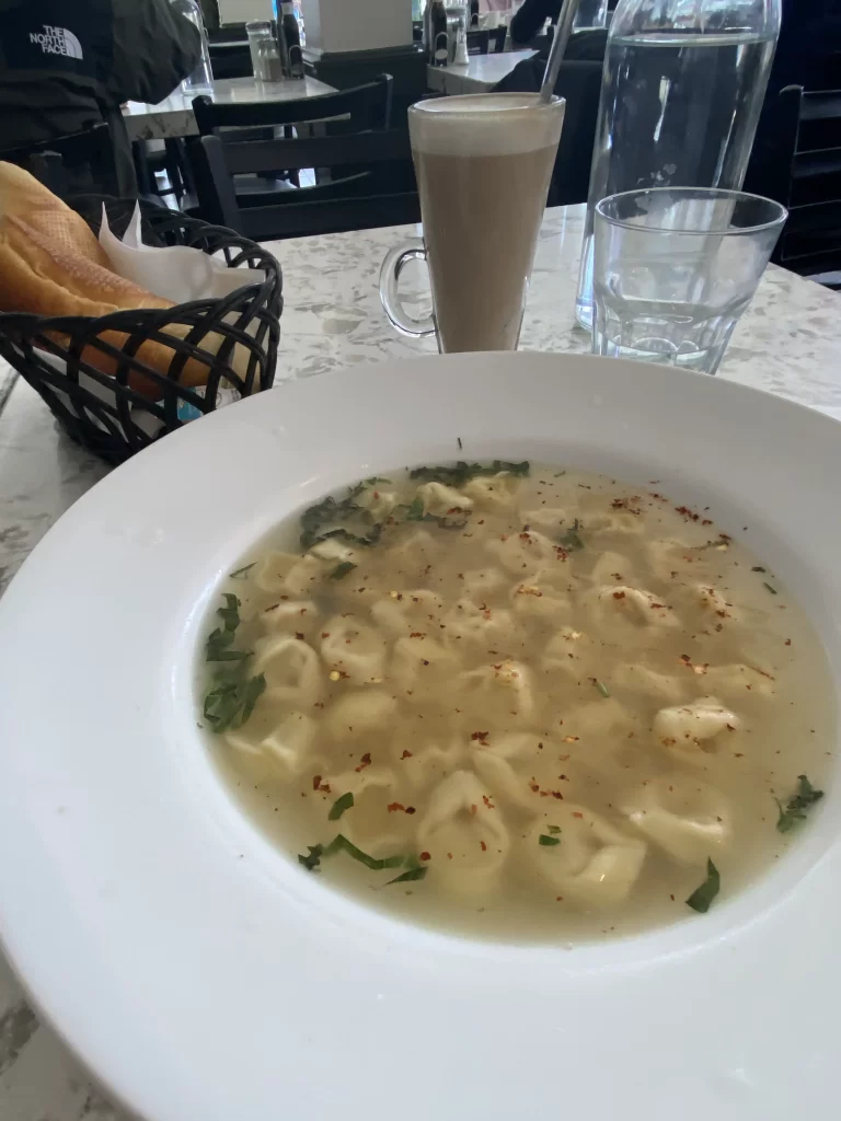 Bowl of tortellini in clear broth garnished with herbs and chili flakes, served with a latte and basket of bread at a marble-topped table inside a bright Toronto restaurant. Large windows offer street views, making it a cozy indoor spot with patio-style light perfect for people watching. A great example of Toronto patio restaurants for people watching.