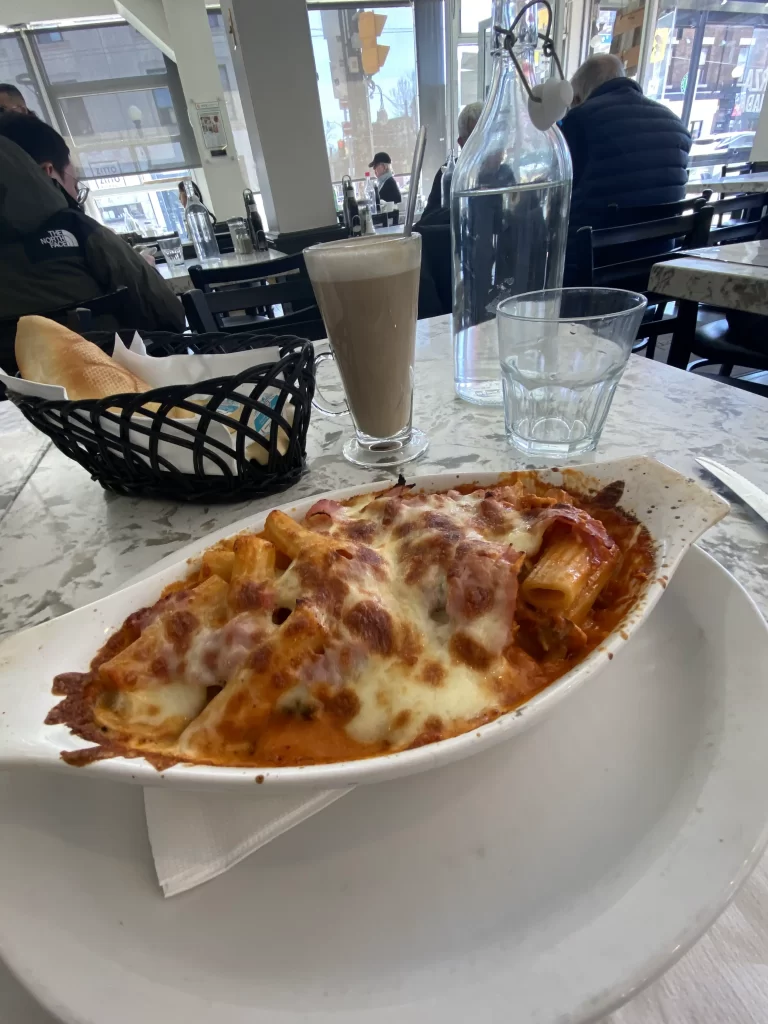 Baked rigatoni covered in melted cheese served at a bright, casual cafe in Toronto with a basket of bread, glass of water, and tall latte on the table. Diners are seated in the background, and the window signage hints at an Italian atmosphere, ideal for enjoying a match. Great example of the cozy, welcoming vibe found in soccer cafes Toronto locals frequent.