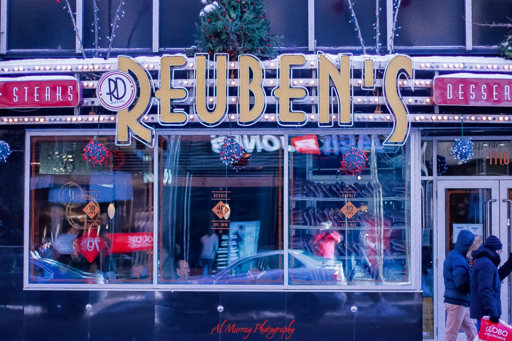 Street view of Reuben’s Deli & Steakhouse with its bold vintage-style sign lit up above large windows decorated for the holidays. Known as one of the best Montreal delis, this family-friendly spot serves classic smoked meat and steakhouse favorites.
