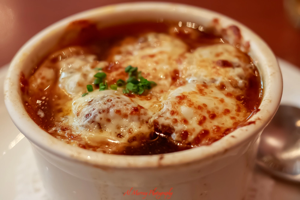 A hot bowl of French onion soup topped with bubbling, golden-brown melted cheese and garnished with chopped green onions. Served as a cozy starter at the family-friendly Reuben’s Deli & Steakhouse in Montreal, known for its smoked meat sandwich.