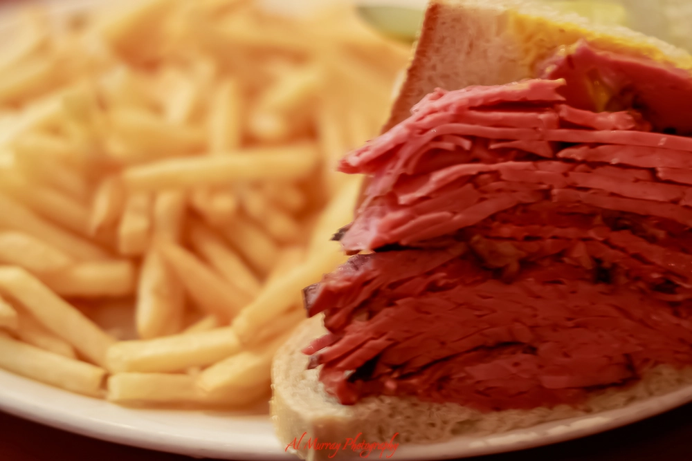 Close-up of a smoked meat sandwich stacked high with thin slices of juicy beef brisket on rye bread, served with a generous side of golden French fries. This classic deli plate highlights the famous smoke meat in Montreal.