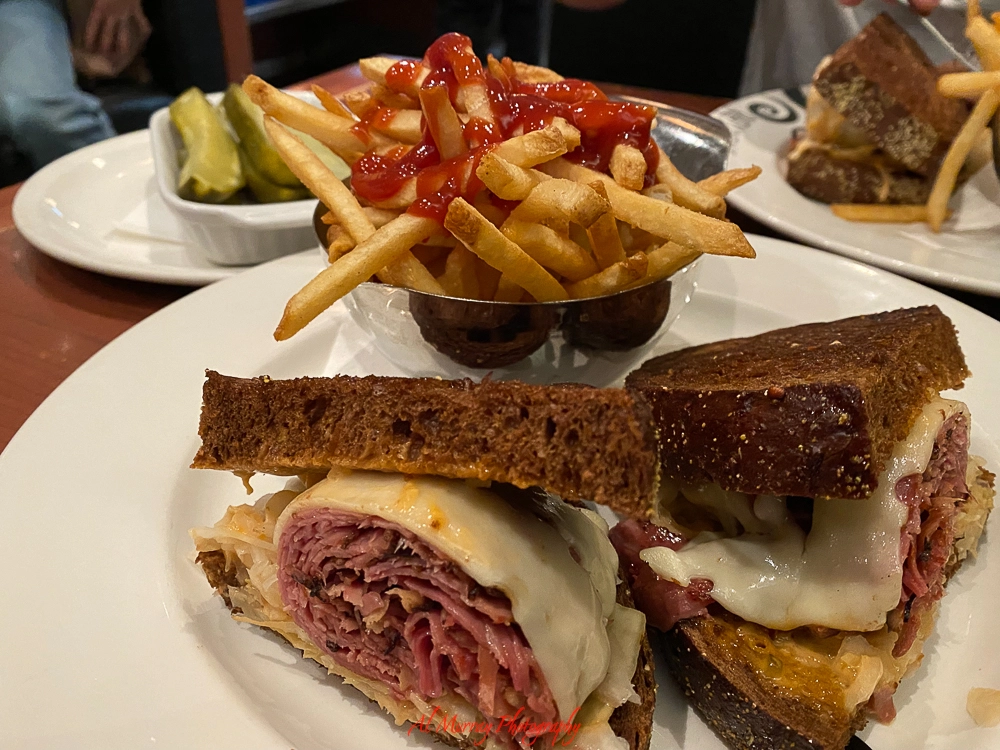 Close-up of a classic Montreal smoked meat sandwich on toasted rye bread, layered with melted Swiss cheese and sauerkraut, served with crispy French fries topped with ketchup and a side of pickles. A hearty example of the famous smoke meat in Montreal.