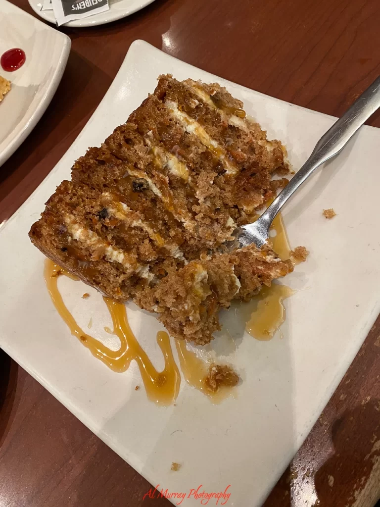 Partially eaten slice of moist carrot cake layered with cream cheese frosting and drizzled with caramel sauce, served on a square white plate. A sweet ending to a quiet early dinner at Reuben’s Deli in Montreal, known for its seniors-friendly atmosphere.