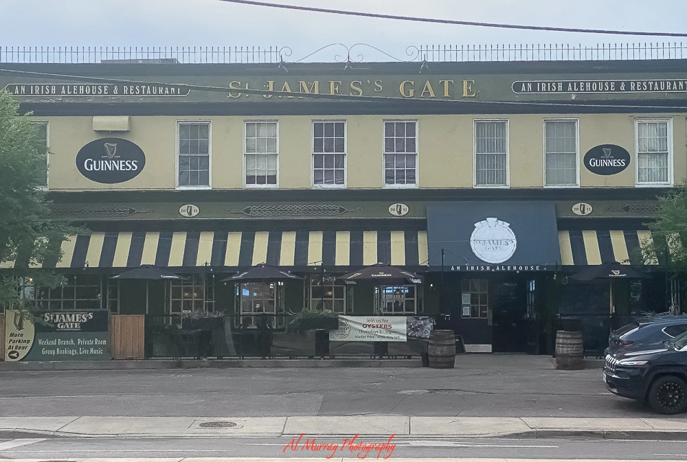 Front view of St. James’s Gate, an Irish pub Etobicoke, with a yellow facade, black-and-yellow striped awnings, and Guinness signs on either side. The patio features black umbrellas, wooden barrels, and a banner promoting oysters on Wednesdays and Sundays. The signage highlights weekend brunch, private rooms, live music, and group bookings.
