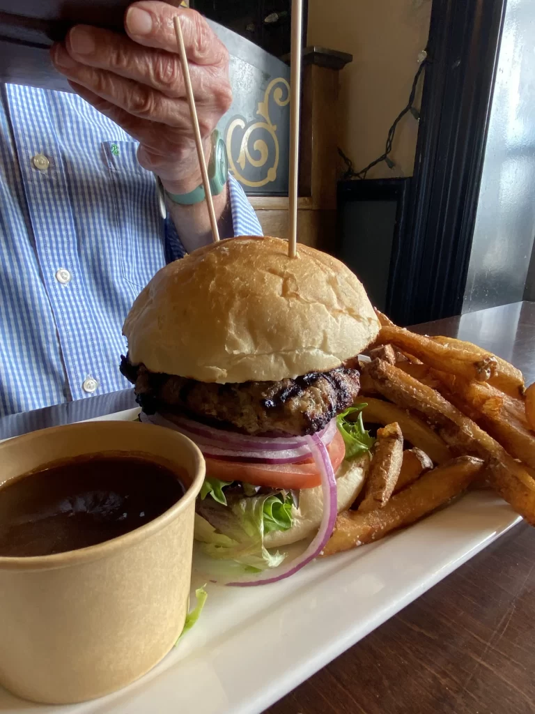 Juicy grilled burger stacked with lettuce, tomato, and red onion on a toasted bun, served with golden fries and a side of gravy at a family friendly pub in Etobicoke. A person in a striped shirt is seen photographing the meal, highlighting a casual dining atmosphere.