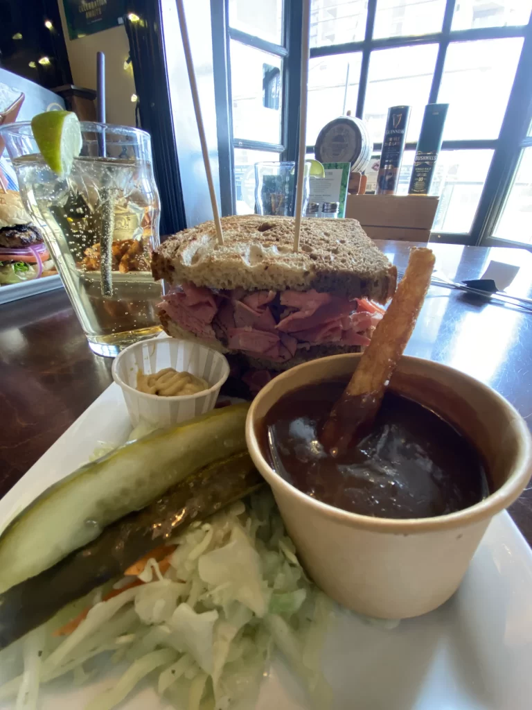 Close-up of a deli sandwich packed with smoked meat on rye bread, served with mustard, a dill pickle spear, coleslaw, and a cup of dark gravy with a fry inside. A glass of cider with lime and beer taps are visible in the background at St. James’s Gate Etobicoke menu, highlighting a hearty option.