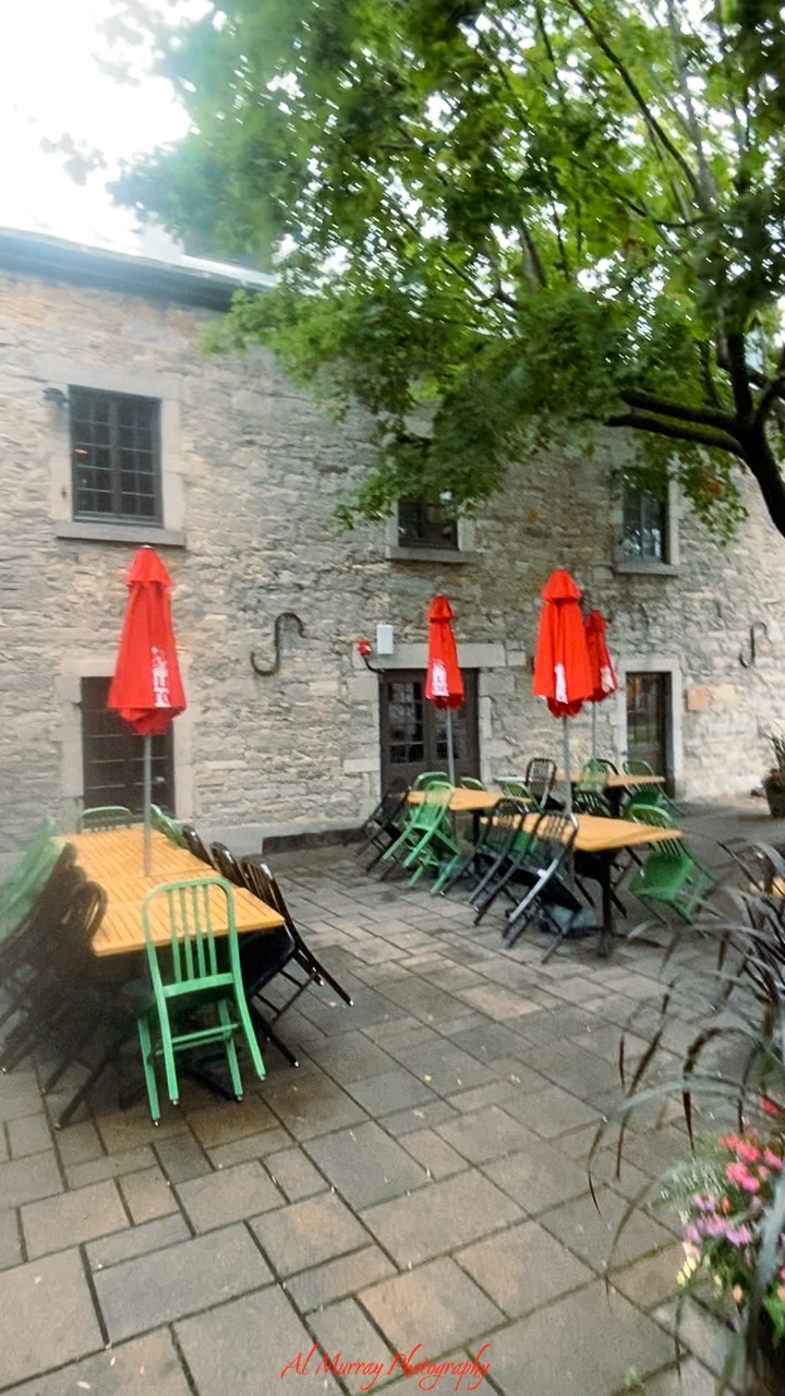 Outdoor patio of a Montreal steakhouse near Old Port, featuring long wooden tables with green and black chairs, shaded by red umbrellas against a rustic stone building. The spacious layout and relaxed setting offer a charming option for open-air dining close to the historic waterfront.