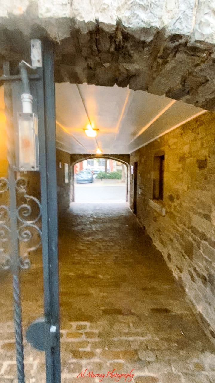 Stone archway entrance leading into a softly lit tunnel with rustic walls and ceiling lights, opening to a view of the street and parked cars beyond. The romantic, historic charm of the passage hints at a hidden gem dining spot, ideal for a "Best steakhouse Old Montreal for couples" experience.