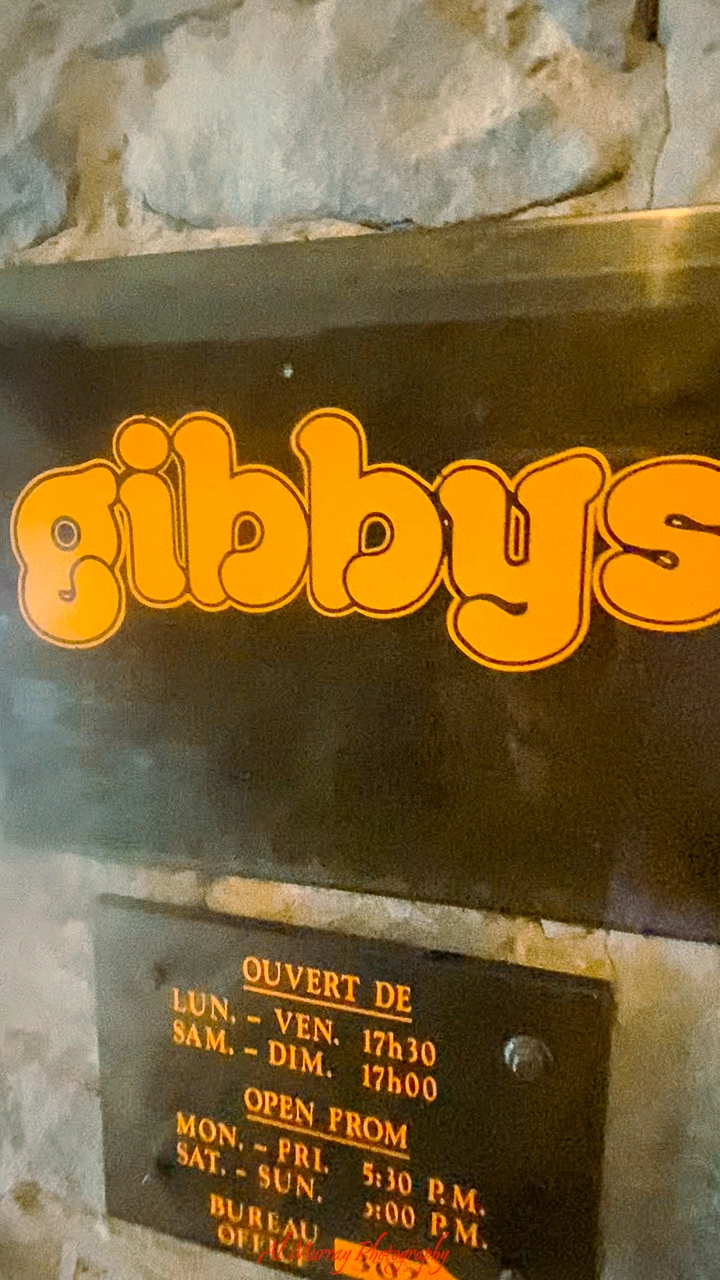 Close-up of the Gibby’s Steakhouse sign featuring bold, retro-style orange lettering against a dark background. Below the name, a plaque lists the restaurant's hours in French, indicating it opens at 5:30 p.m. on weekdays and weekends, useful for planning an early dinner in a Gibby’s Steakhouse Old Montreal review for seniors.