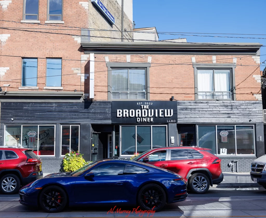 Street view of The Broadview Diner, a modern black-and-brick storefront with large windows and a visible "OPEN" sign, located in a walkable area near Broadview station. Parked cars line the street, emphasizing easy access to one of several walkable restaurants near Broadview station.