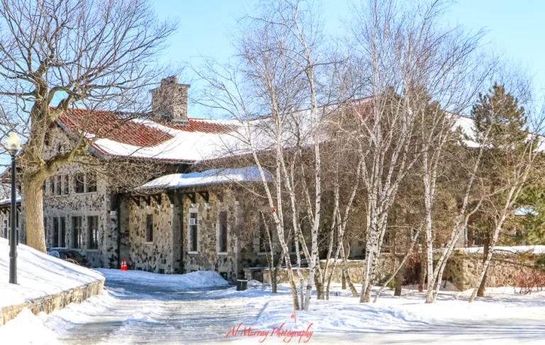 A rustic stone building with a red-tiled roof sits nestled among bare trees and snow-covered ground under a clear blue sky in Montreal. This cozy winter scene reflects the city’s historic charm and highlights the importance of seasonal awareness in any list of 9 Montreal travel tips.