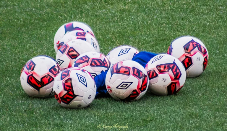 Close-up of a cluster of white and red Umbro soccer balls resting on green turf, with a blue training vest tucked between them. A vibrant and energetic scene symbolizing the excitement building as fans prepare to visit Vancouver for World Cup 2026.