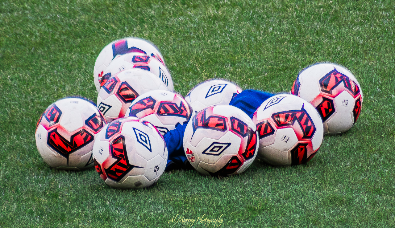 Close-up of a cluster of white and red Umbro soccer balls resting on green turf, with a blue training vest tucked between them. A vibrant and energetic scene symbolizing the excitement building as fans prepare to visit Vancouver for World Cup 2026.