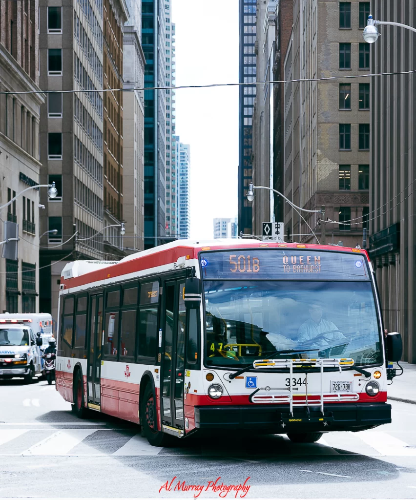 how to save money on toronto world cup 2026 hotels A red and white TTC bus labeled 501B Queen to Bathurst drives through a downtown Toronto intersection surrounded by tall buildings. Using public transit like this is a smart way for World Cup 2026 visitors to save money on accommodation by staying in more affordable areas outside the city core.