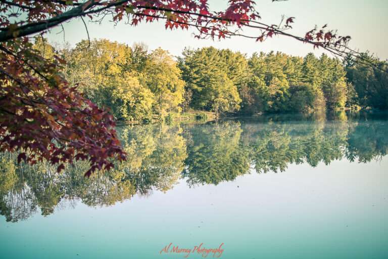 Calm lake reflecting vibrant autumn trees with red maple leaves framing the scene in the foreground. This tranquil setting offers a peaceful nature experience ideal for accessible travel in Canada for seniors.