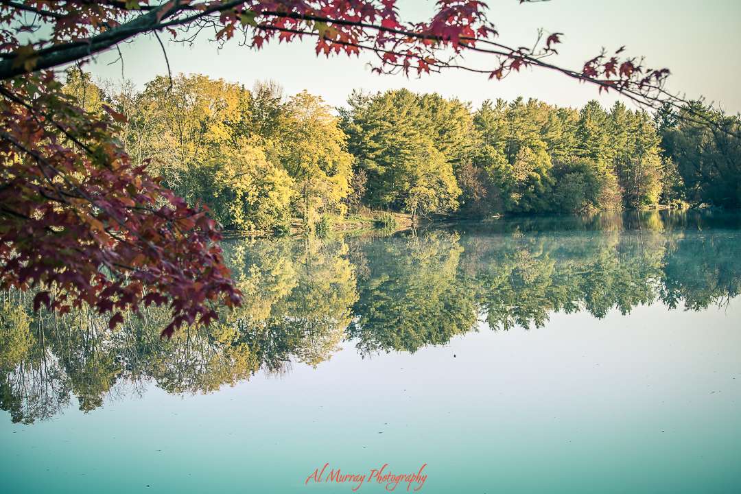 Calm lake reflecting vibrant autumn trees with red maple leaves framing the scene in the foreground. This tranquil setting offers a peaceful nature experience ideal for accessible travel in Canada for seniors.