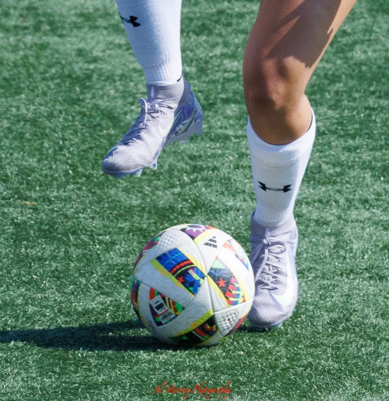 Close-up of a soccer player in white Under Armour socks and cleats controlling a colorful match ball on a turf field. The dynamic action highlights soccer culture, relevant for a World Cup 2026 Canada travel guide