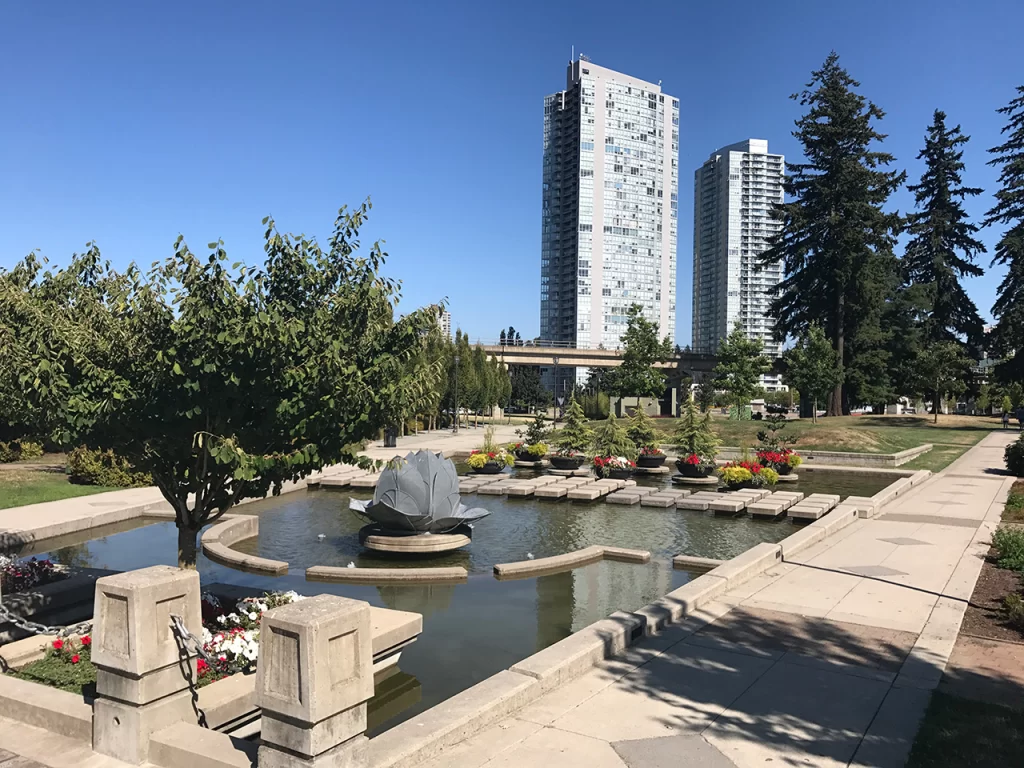 Vancouver budget stays suburbs Sunny day at a landscaped urban park in Metro Vancouver, featuring a lotus-shaped fountain, colorful flowerbeds, and high-rise apartment buildings in the background. Peaceful spots like this are often steps away when you find budget accommodation in Vancouver area suburbs.