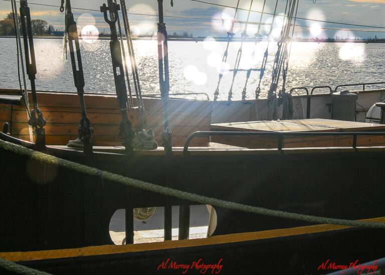Close-up of a wooden sailing ship docked at a harbor during sunset in Toronto, with glowing sunlight reflecting on the water and lens flares across the image. The peaceful waterfront scene hints at budget-friendly travel moments, tying into how to save money on accommodation for World Cup 2026 in Toronto.