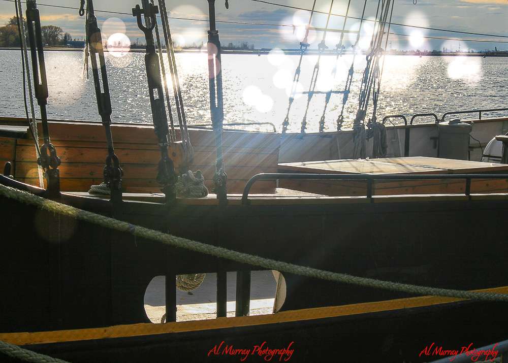 Close-up of a wooden sailing ship docked at a harbor during sunset in Toronto, with glowing sunlight reflecting on the water and lens flares across the image. The peaceful waterfront scene hints at budget-friendly travel moments, tying into how to save money on accommodation for World Cup 2026 in Toronto.