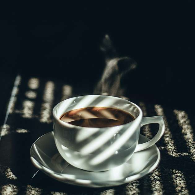 White ceramic cup filled with steaming coffee, resting on a matching saucer in dramatic lighting with shadows from window blinds. The moody, cozy setting evokes the warm atmosphere often found in the best local coffee shops in Toronto.