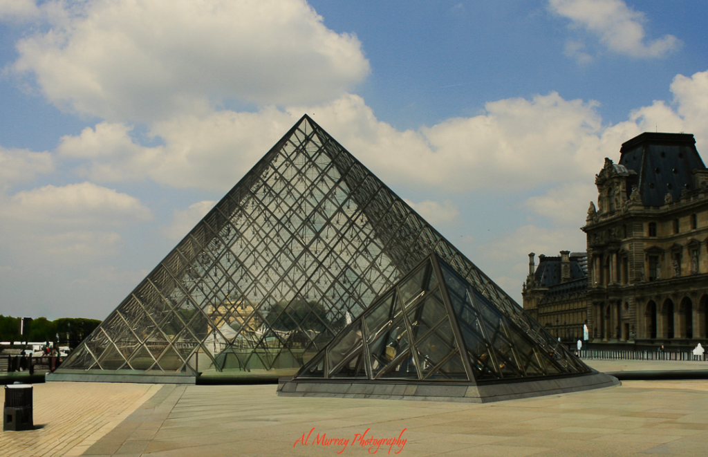 The Louvre Pyramid in Paris, a striking glass and metal structure with classical French architecture in the background under a partly cloudy sky. A popular cultural stop for tennis fans visiting the French Open, especially older travelers seeking enriching off-court experiences.