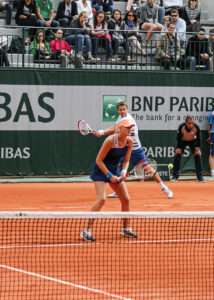 Two tennis players compete in a doubles match on a clay court at the French Open, with a packed audience watching from the stands. The male player in the background is mid-swing, while the female player in the foreground is positioned to return the ball, highlighting the excitement of live matches for senior tennis fans visiting Roland-Garros.
