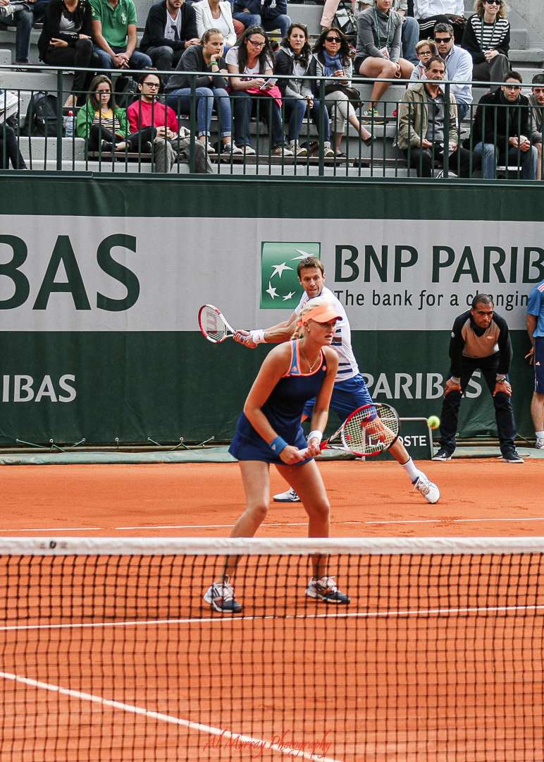 Two tennis players compete in a doubles match on a clay court at the French Open, with a packed audience watching from the stands. The male player in the background is mid-swing, while the female player in the foreground is positioned to return the ball, highlighting the excitement of live matches for senior tennis fans visiting Roland-Garros.