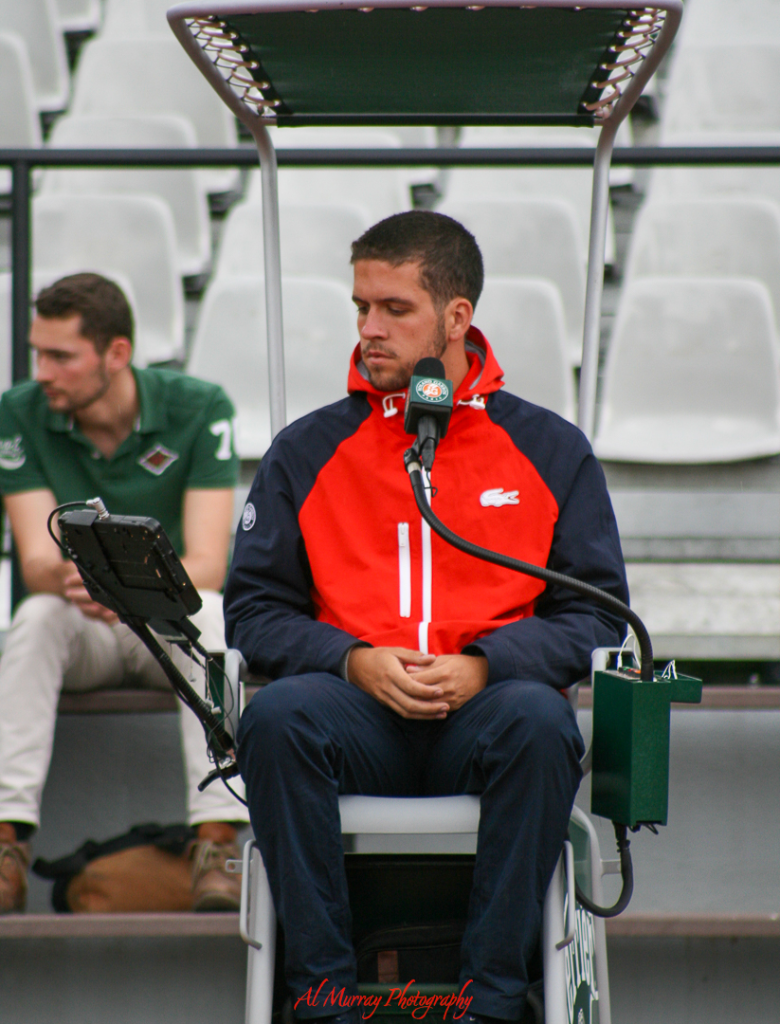 Tennis chair umpire in a red and navy jacket seated on a high umpire chair, focused and ready during a match at a professional stadium with empty white seats in the background. This scene reflects the formal officiating seen at international tennis tournaments, important for seniors planning travel to major events.