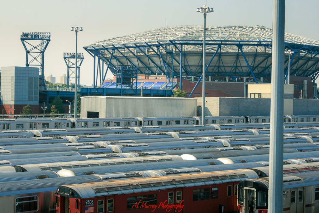US Open travel guide for older tennis fans Rows of MTA subway trains are parked in a rail yard near Arthur Ashe Stadium in New York City, with the stadium’s blue steel frame and seating visible in the background. This view highlights the accessibility of public transit for visitors using a US Open travel guide for older tennis fans